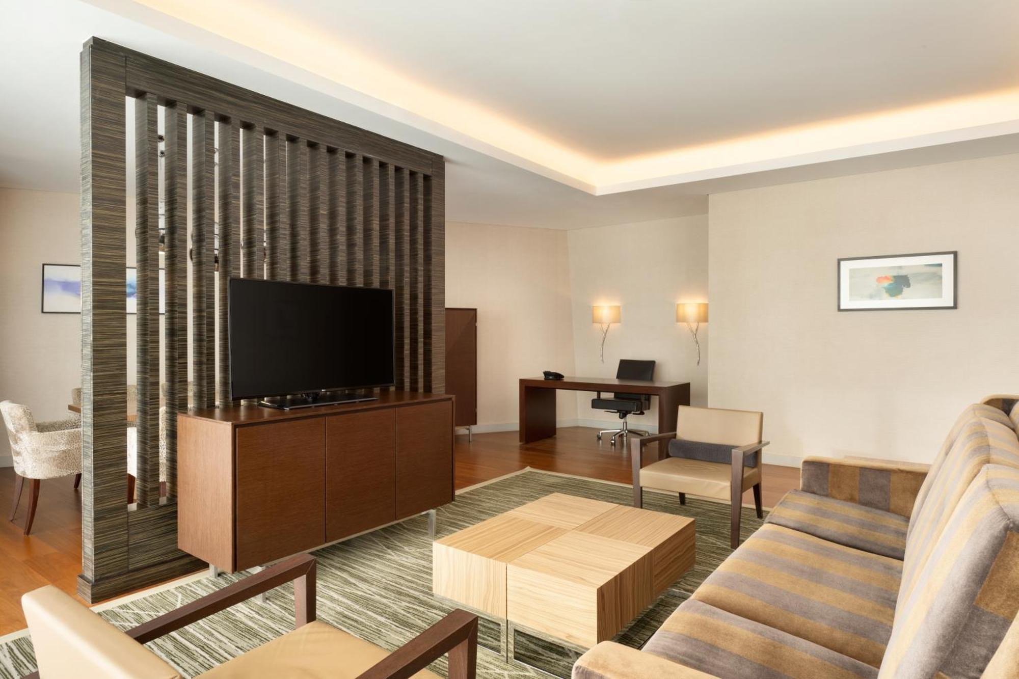 Hilton Riyadh Hotel & Residences Exterior foto The photo shows a modern living space that features a contemporary design. It includes a seating area with a beige sofa and two armchairs arranged around a square wooden coffee table. A wall-mounted flat-screen TV is positioned on a decorative wooden